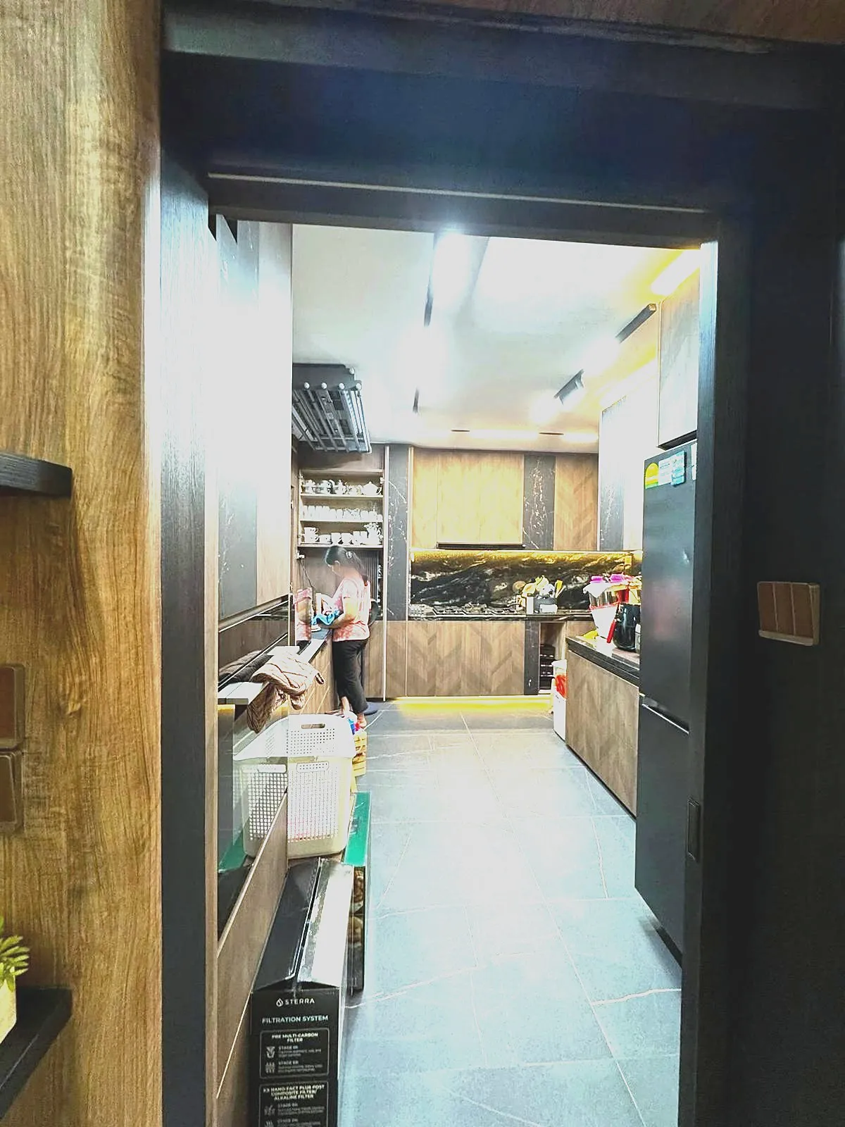 A galley kitchen in dark timber veneer with chevron-laid cabinet fronts, a black marble backsplash and under-cabinet strip lighting