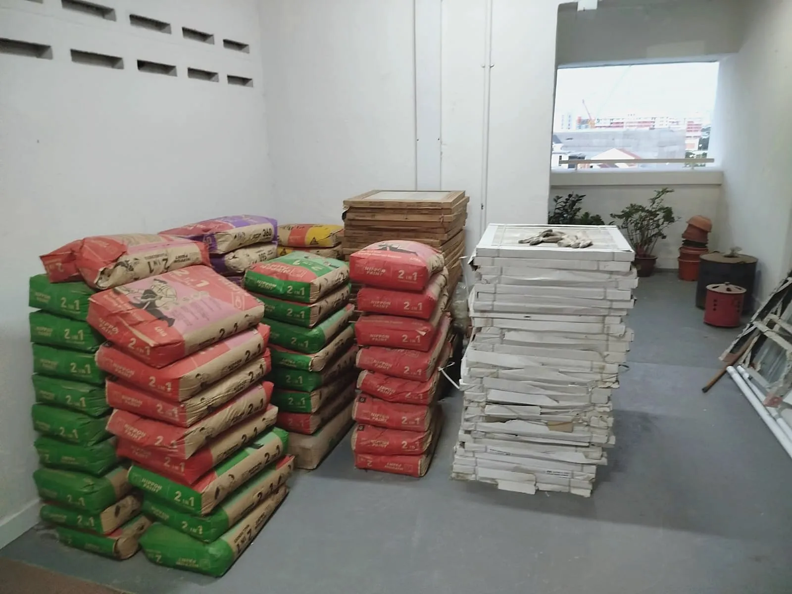 Pallets of skim coat and cement stacked against a wall of a site store, beside a stack of boxed floor tiles