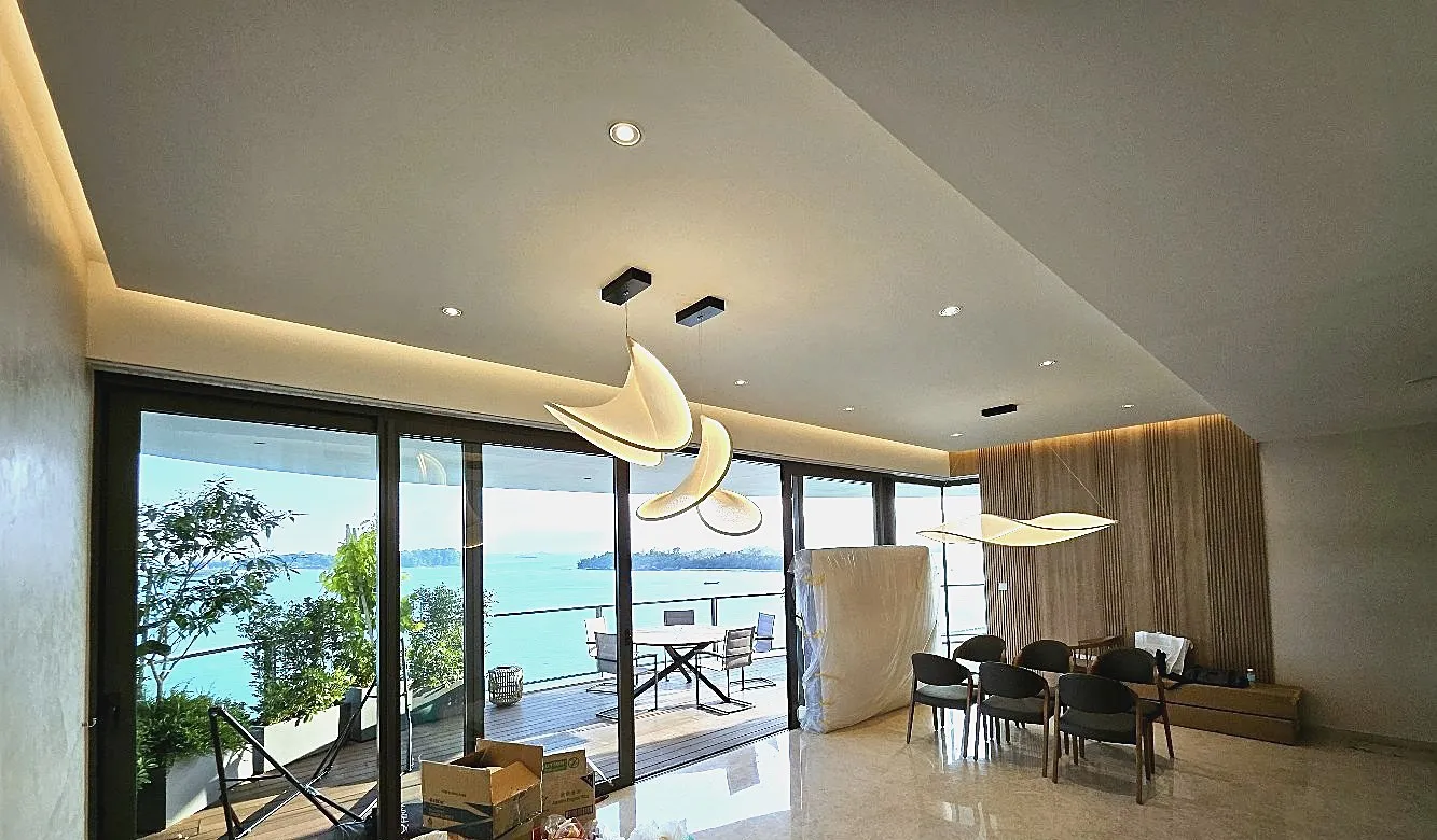 A completed living and dining room with timber joinery, pendant lighting over the table and a full-height window looking out to the sea